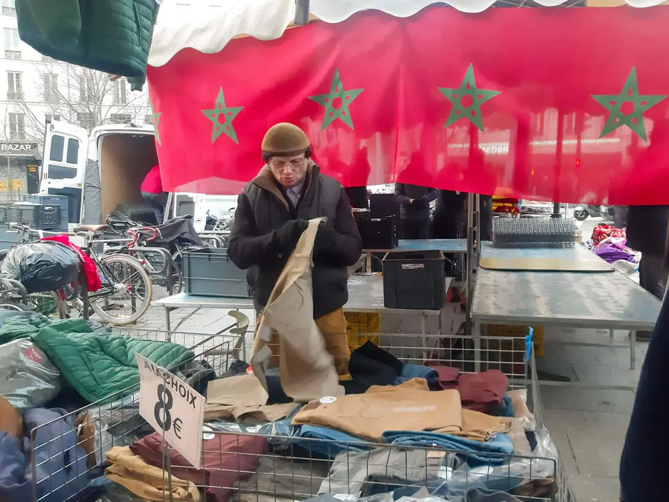 Un cliente escoge una ropa con una bandera de Marruecos de fondo en el mercado de Belleville en París este martes. En el de Belleville, el mercado más árabe de París, se dan cita marroquíes, egipcios, tunecinos y argelinos que apoyan a Marruecos, que se enfrentará mañana a Francia en un partido de semifinales del Mundial de Fútbol Qatar 2022. EFE/ Antonio Torres Del Cerro